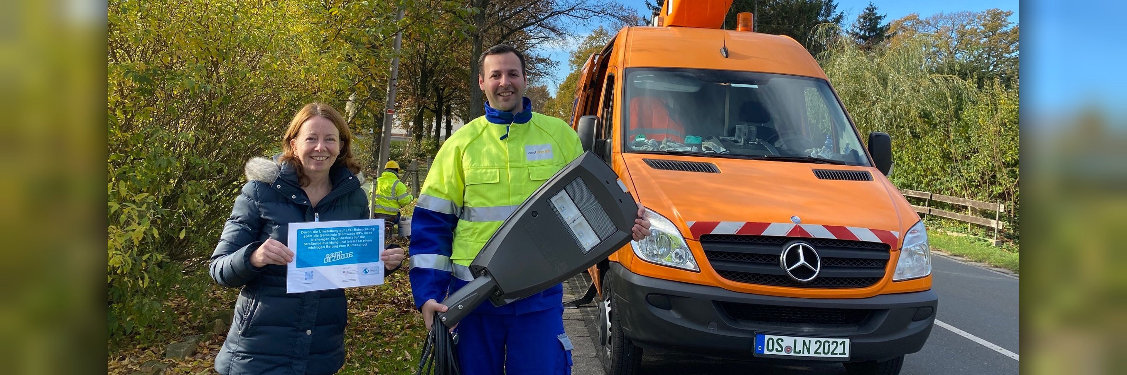 Eine Frau und ein Mann stehen mit einer neuen LED-Leuchte vor einem Kranfahrzeug.