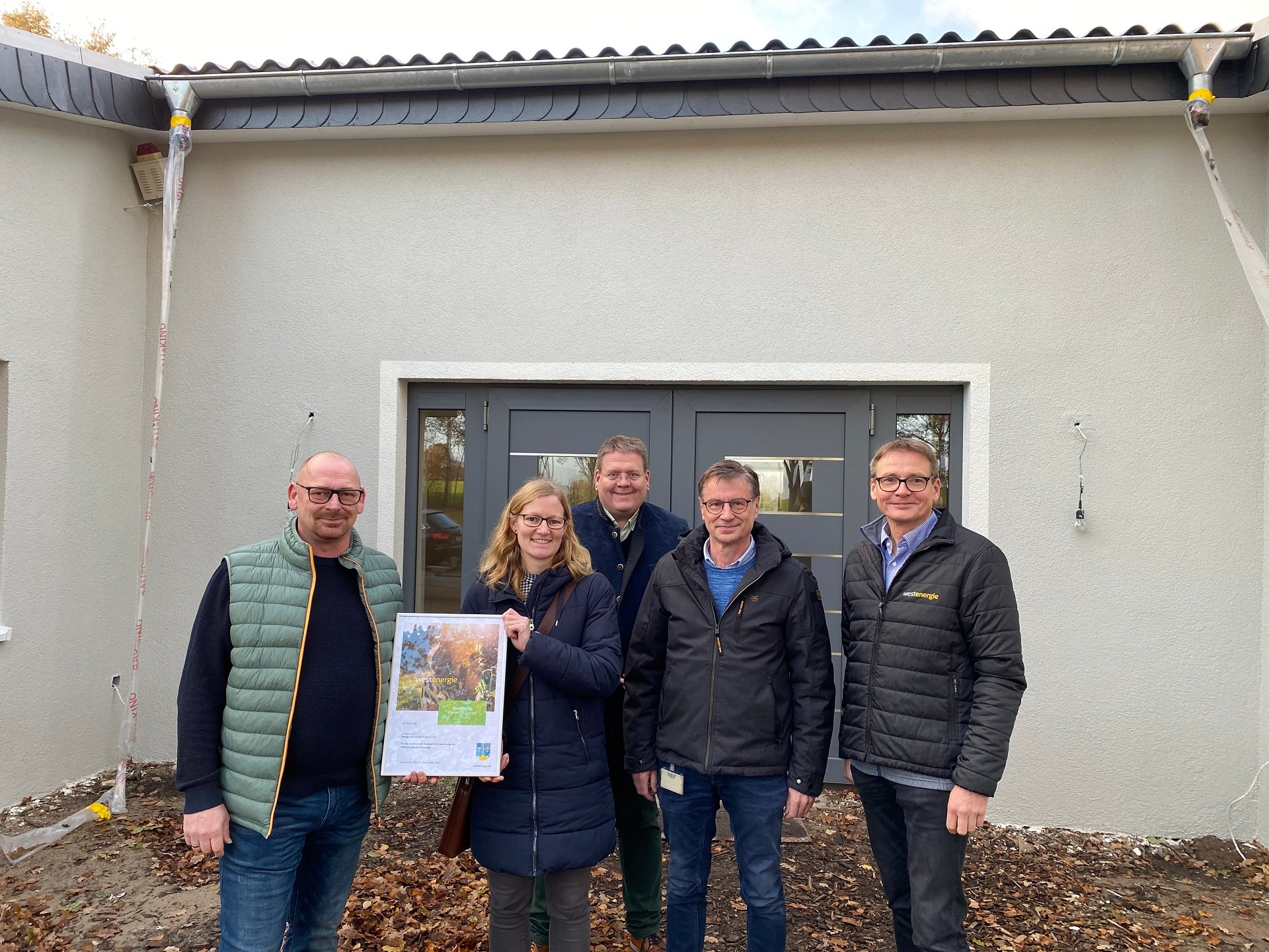 Eine Gruppe von Frauen und Männern, die vor einem noch nicht ganz fertig gebauten Haus steht und eine Urkunde in die Kamera hält. 