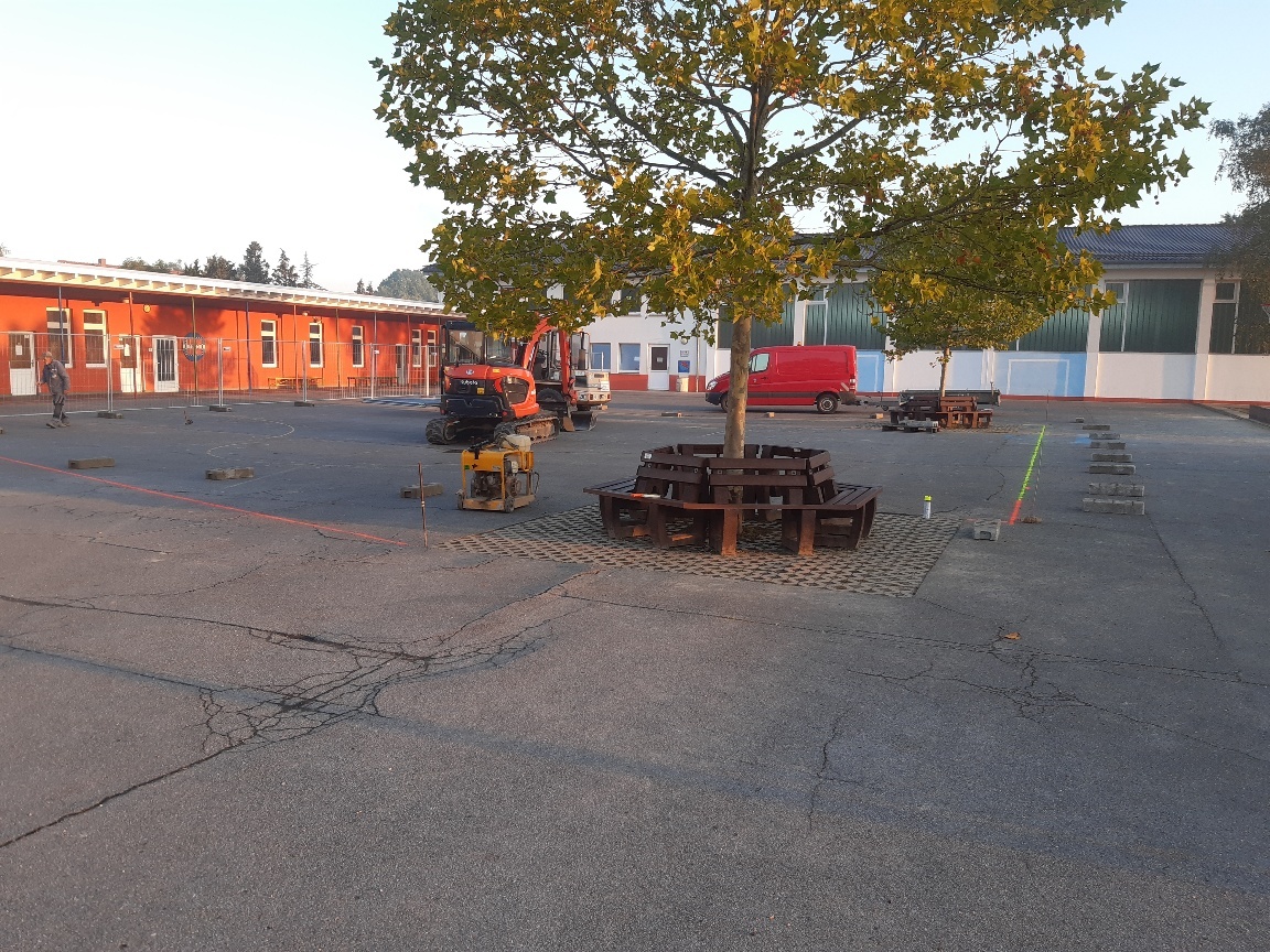 Ein Bagger steht auf einem asphaltierten Schulhof. Ein Arbeiter malt Markierungen auf den Boden. 