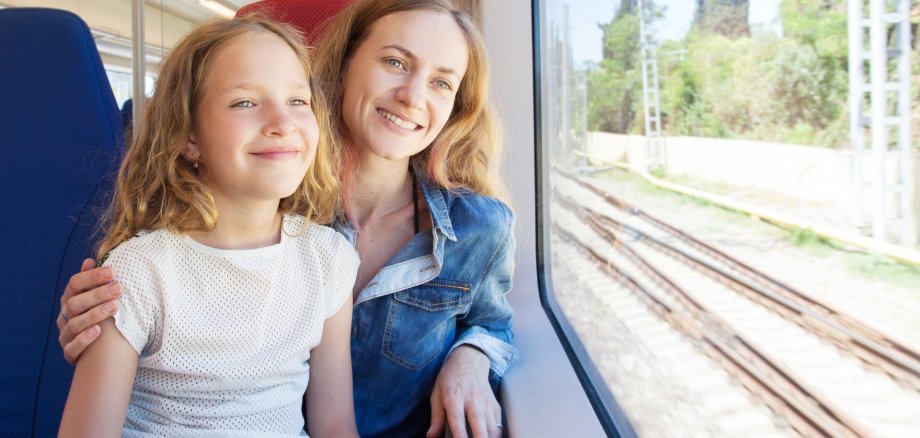 Eine junge Mutter und ihre Tochter sitzen in einem fahrenden Zug und schauen gut gelaunt aus dem Fenster.