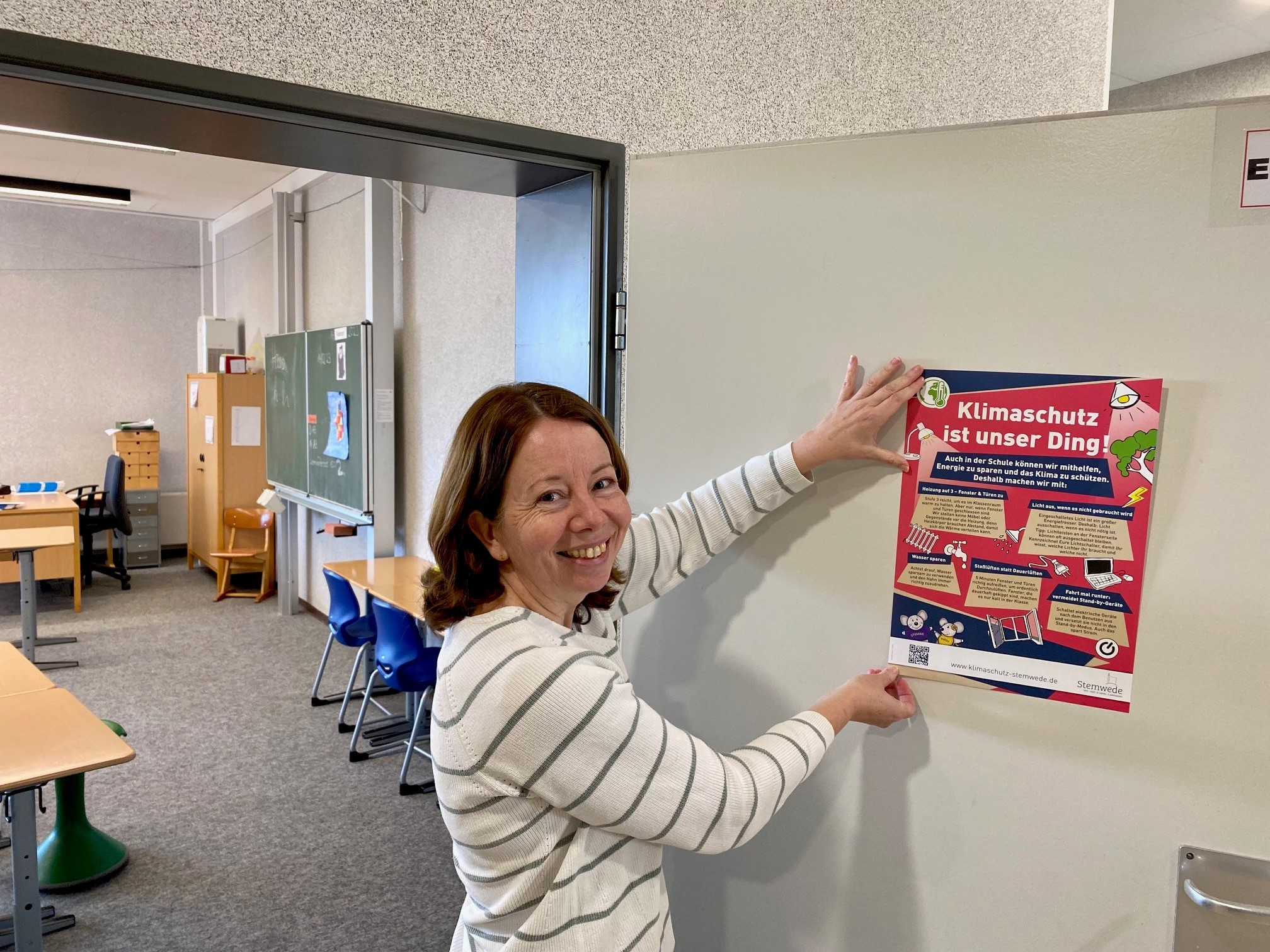 Meldung Energiesparplakate Eine Frau steht vor einem Klassenraum und klebt ein Plakat mit Energiespartipps an die Tür.