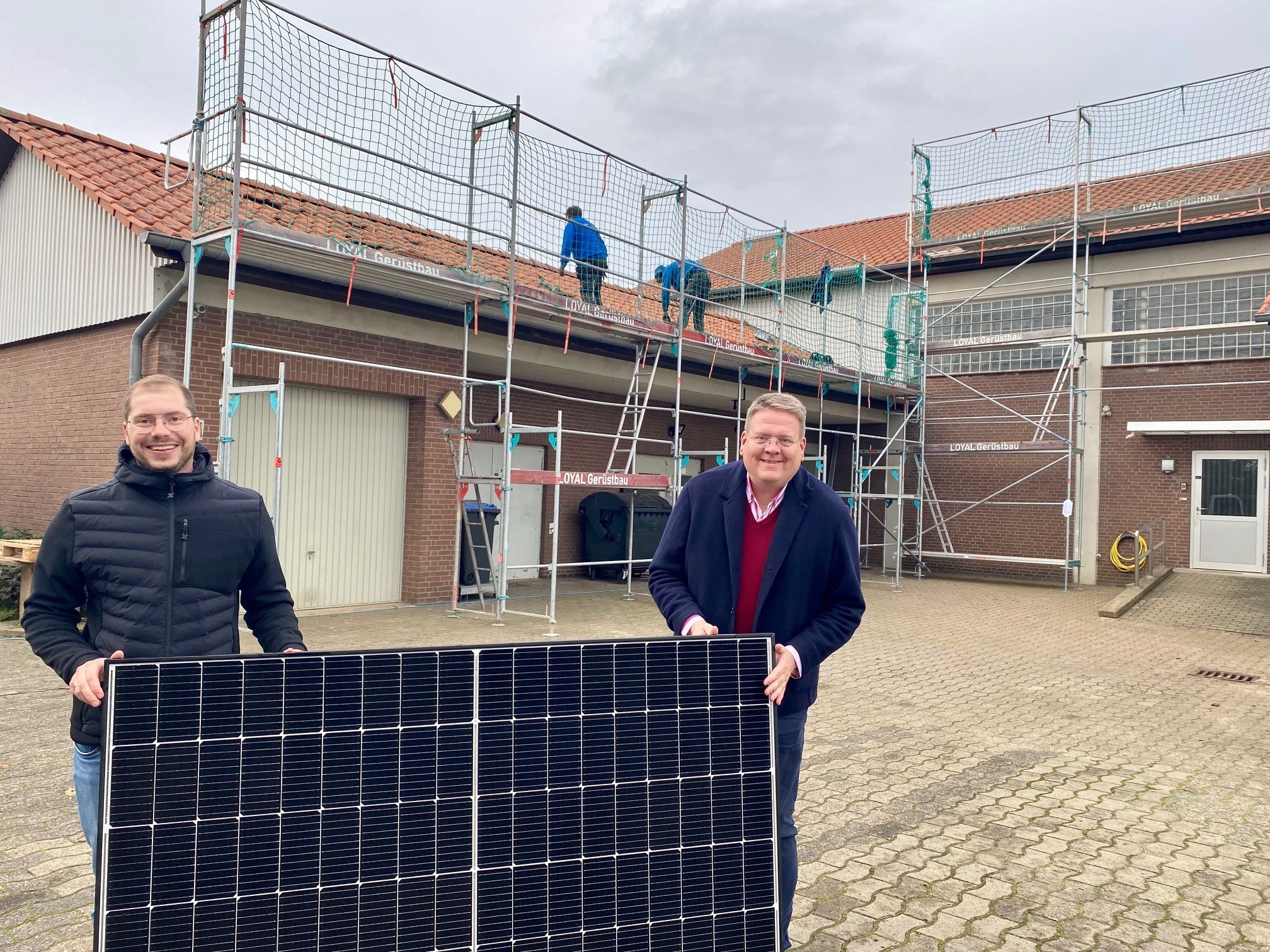 Zwei Männer halten ein Solarmodul. Im Hintergrund ist ein rotes Gebäude zu sehen, dass mit einem Baugerüst umstelt ist. 