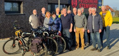 Eine Gruppe Radfahrerinnen und Rafahrer posiert für ein Gruppenfoto vor einem Feuerwehrgerätehaus. 