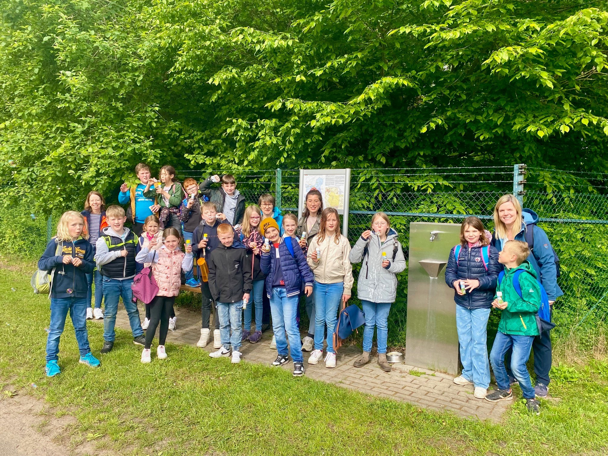 Eine Gruppe von Kindern, die für ein Gruppenfoto an einem Trinkwasserspender posieren. Die Kinder haben Rucksäcke auf dem Rücken und viele von ihnen haben Seifenblasen in der Hand.