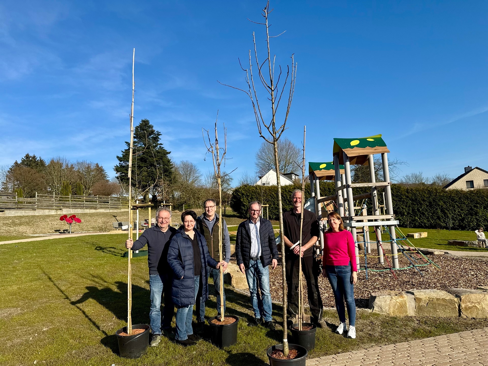 Sechs Personen haben sich vor einem Klettergerüst auf einem Spielplatz positioniert und präsentieren mehrere zum Einpflanzen bereite Bäume.