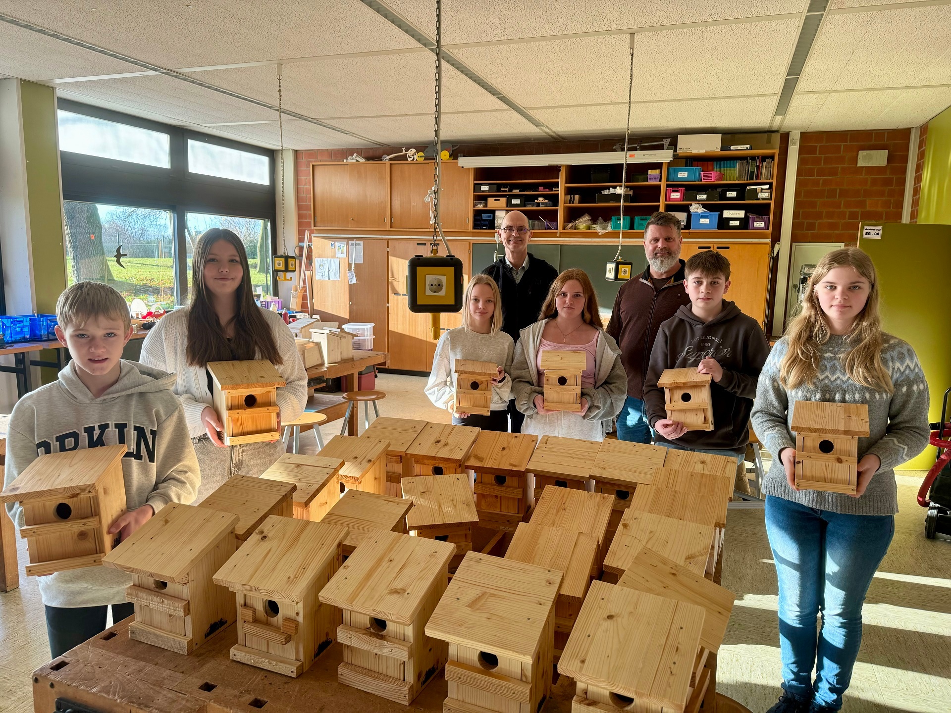 Eine Gruppe von Schülerinnen und Schülern steht, zusammen mit zwei erwachsenen Männern, in einem Werkraum einer Schule, in dem zahlreiche hölzernen Vogelhäuser auf den Werkbänken stehen und schauen in die Kamera.