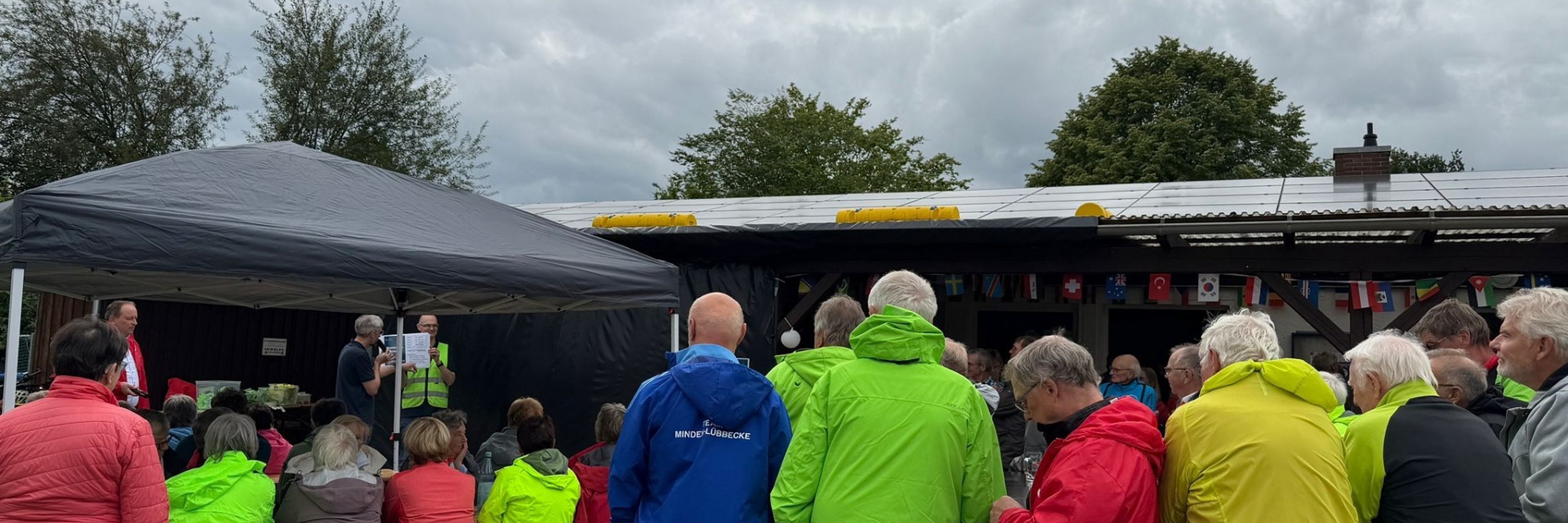 STADTRADELN-Abschluss 2026 Teilnehmerinnen und Teilnehmer des STADTRADELN-Abschlusstages versammeln sich vor einem Pavillon auf dem Sportplatz in Levern und verfolgen eine Ansprache. Viele Gäste tragen wetterfeste Jacken in leuchtenden Farben und sitzen oder stehen dicht beieinander. Im Hintergrund sind internationale Flaggen zu sehen, während dunkle Regenwolken über dem Veranstaltungsgelände hängen.