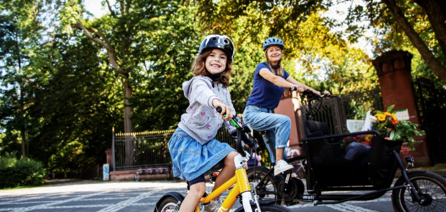 Ein Kind mit Fahrradhelm fährt auf einem gelben Kinderfahrrad im Vordergrund und lächelt in die Kamera. Dahinter fährt eine erwachsene Person mit Helm auf einem Lastenfahrrad. Beide befinden sich auf einer ruhigen, von Bäumen gesäumten Straße bei sonnigem Wetter. Im Lastenrad sind Taschen und Blumen zu sehen.