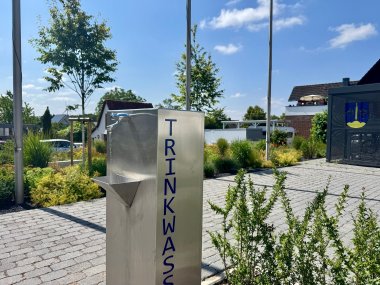 Trinkwasserspender Stemwede Edelstahl-Trinkwasserspender mit der vertikalen Aufschrift ‚TRINKWASSER‘ auf einem gepflasterten Platz. Im Hintergrund befinden sich Bäume, bepflanzte Grünflächen, Fahnenmasten sowie Wohnhäuser unter einem sonnigen, blauen Himmel mit wenigen Wolken.